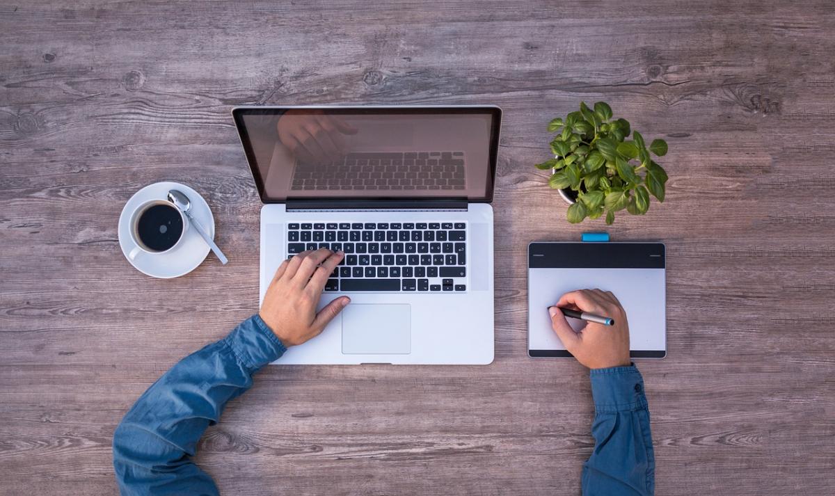 guy in a blue shirt sitting at a laptop with a cup of coffee, a drawing pad and a plant